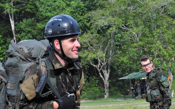 Two soldiers in camouflage uniforms are visible in an outdoor field setting surrounded by trees. The foreground soldier is fully equipped with a large rucksack, tactical gear, helmet, gloves, and carries a rifle marked with the number '10'. The background soldier wears protective eyewear and similar camouflage attire, standing at ease.