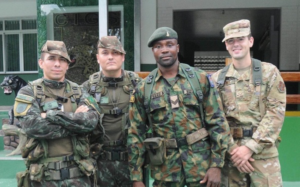 Four military personnel in different uniforms pose together outdoors on a green surface in front of a building with partial text 'CIGS' visible. Three appear to wear Brazilian Army uniforms with distinctive woodland camouflage patterns, while the fourth (rightmost) wears US military MultiCam/OCP pattern uniform. The image appears to document a multinational military exchange or joint training event.