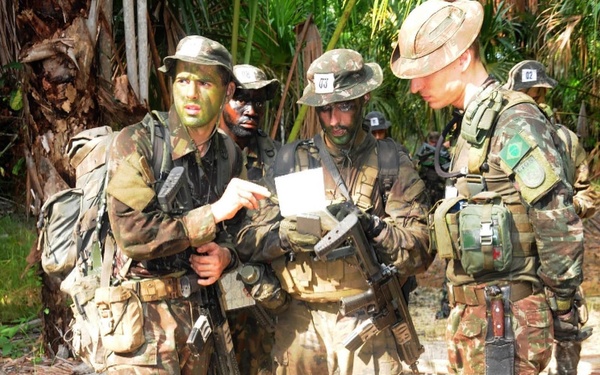 A group of military personnel in jungle/tropical environment wearing camouflage uniforms and face paint, appearing to be engaged in a field exercise or patrol operation. The soldiers are equipped with full combat gear including weapons, tactical vests, and backpacks. A Brazilian Army flag patch is visible on at least one soldier's uniform, suggesting this may be a Brazilian military exercise.