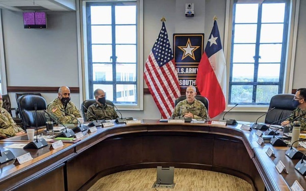 A formal military briefing or command meeting is taking place at U.S. Army South headquarters, with approximately 12-15 uniformed personnel seated around a large U-shaped conference table. A senior officer appears to be presiding at the head of the table, with the U.S. flag and Texas state flag flanking a U.S. Army South emblem in the background. Several attendees are wearing face masks, suggesting this was taken during the COVID-19 pandemic era.