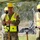 Three military personnel in camouflage uniforms are wearing yellow safety vests and hard hats, standing at what appears to be a groundbreaking ceremony. The central figure appears to be speaking or presenting, while the others stand at attention. One vest is labeled '919th SOW', indicating affiliation with the 919th Special Operations Wing.