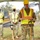 A group of military personnel in camouflage uniforms are participating in a groundbreaking ceremony at a construction site. The central figure, wearing a high-visibility safety vest and yellow hard hat, appears to be speaking or presenting, while three others stand behind him also wearing safety vests and hard hats. Three of the four background personnel are wearing black face masks, suggesting this event took place during the COVID-19 pandemic period.