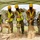 A groundbreaking ceremony is taking place at a military or government construction site, with five individuals simultaneously shoveling dirt with ceremonial gold-painted shovels. Four participants are wearing military camouflage uniforms (OCP pattern) with high-visibility safety vests and yellow hard hats, while one civilian wears a white hard hat with a logo and a safety vest. All participants are wearing black face masks, suggesting the event occurred during the COVID-19 pandemic period.
