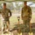 Two U.S. Army soldiers in OCP (Operational Camouflage Pattern) uniforms stand under a green canopy tent at a military groundbreaking ceremony. They are posed with ceremonial gold-painted shovels planted in the ground, with construction equipment including a John Deere excavator visible in the background. The setting appears to be an outdoor construction site on a military installation.