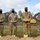 Four military personnel in OCP (Operational Camouflage Pattern) uniforms and black face masks pose with ceremonial gold shovels at what appears to be a groundbreaking ceremony. They are standing in front of a mound of disturbed earth with construction equipment visible in the background. A helicopter is faintly visible in the upper right portion of the sky.