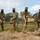 Four military personnel in OCP (Operational Camouflage Pattern) uniforms are participating in a groundbreaking ceremony, using ceremonial gold-painted shovels to turn dirt. All four individuals are wearing black face masks, suggesting the event took place during the COVID-19 pandemic period. Construction equipment, including a John Deere bulldozer and a CAT 135G excavator, is visible in the background behind a chain-link fence.