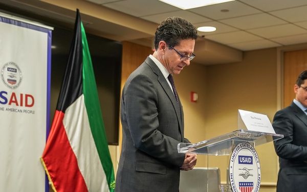 Two men in business suits stand at clear acrylic podiums bearing the USAID (United States Agency for International Development) seal during what appears to be an official signing ceremony or press event. The setting includes a USAID banner, an American flag, and what appears to be a Kuwaiti flag in the background. The primary speaker appears to be reviewing notes at the podium while a second official stands at a separate podium to the right.