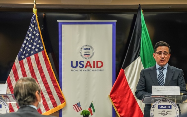 A formal diplomatic or partnership event hosted by USAID (United States Agency for International Development) featuring representatives from the United States and Kuwait. A speaker stands at a podium bearing the USAID seal, with the American and Kuwaiti flags prominently displayed alongside a USAID banner. The setting suggests an official signing ceremony or press event, with attendees seated in a socially distanced arrangement consistent with COVID-19 protocols.