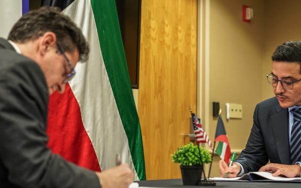 Two men in dark suits are seated at a black-draped table signing documents in what appears to be a formal agreement or treaty signing ceremony. A US flag and what appears to be a Kuwait flag are visible in the background, along with a USAID banner. A third man wearing a face mask stands in the background observing the proceedings.