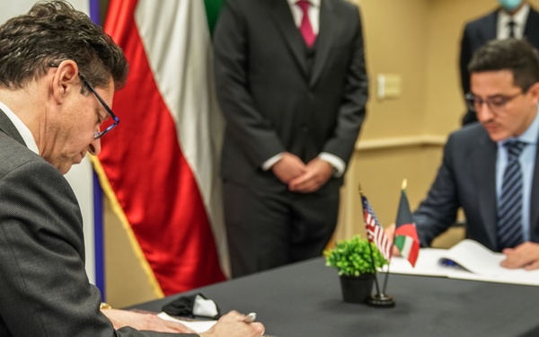 Two men in business suits are seated at a dark-draped table engaged in a document signing ceremony. Small desktop flags, including a US flag and what appears to be an Afghan flag, are visible on the table between them. A third person stands in the background near several larger flags, and a fourth individual wearing a face mask is partially visible in the far background.