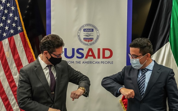 Two officials in business suits and face masks perform an elbow bump greeting at a signing ceremony table in front of a USAID banner. The setting includes both an American flag and what appears to be a Kuwaiti flag, along with small desk flags of both nations, suggesting a bilateral agreement or partnership event. Documents are visible on the table, indicating a formal signing has taken or is about to take place.