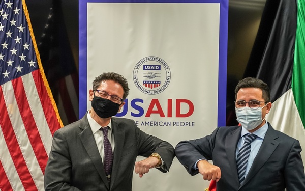 Two officials wearing face masks perform an elbow bump greeting at a USAID signing ceremony. They are standing in front of a USAID banner flanked by an American flag and what appears to be a Kuwaiti flag, with signed documents visible on the table before them. The setting suggests a formal bilateral agreement or partnership signing between the United States and Kuwait.