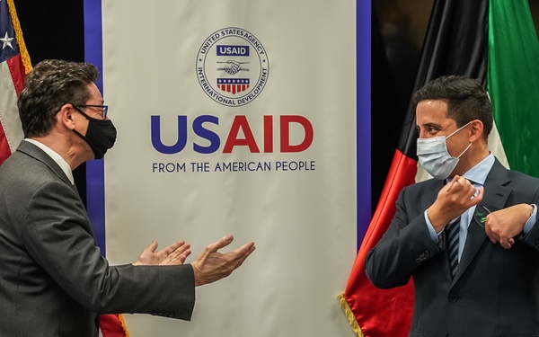 Two masked individuals are engaged in conversation at what appears to be an official signing ceremony hosted by USAID (United States Agency for International Development). The setting includes national flags and a signing table with documents, suggesting a formal bilateral agreement or partnership event. Both individuals are wearing face masks, consistent with COVID-19 era protocols.