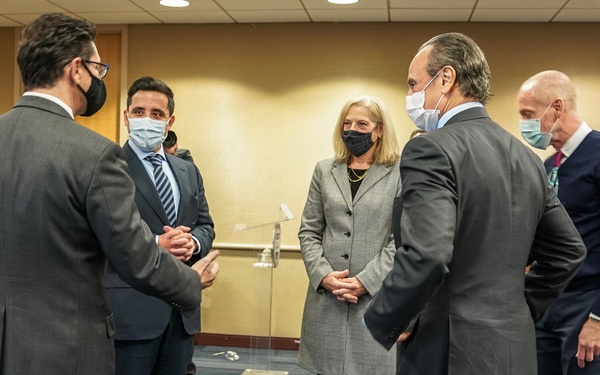 A group of professionally dressed individuals are gathered in what appears to be a conference or meeting room, engaged in conversation. All visible participants are wearing face masks, suggesting this was taken during the COVID-19 pandemic period. The setting appears to be a formal or semi-formal networking or post-meeting gathering.
