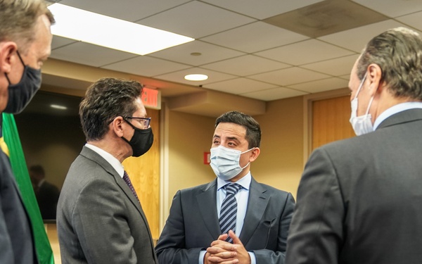 A group of professionally dressed individuals in business suits are gathered in what appears to be a conference or meeting room, engaged in conversation. All visible participants are wearing face masks, suggesting this was taken during the COVID-19 pandemic period. The central figure appears to be speaking to the group with hands clasped together.