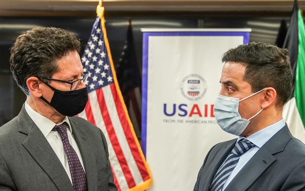 Two men in business suits wearing face masks are engaged in conversation at what appears to be an official USAID event or meeting. The setting includes an American flag and what appears to be a UAE (United Arab Emirates) flag in the background, along with a USAID banner. The context suggests a formal diplomatic or aid-related engagement.