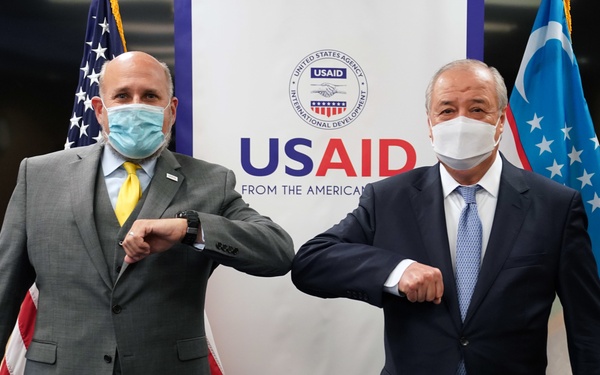 Two men wearing face masks are performing an elbow bump greeting in front of a USAID (United States Agency for International Development) banner. The setting appears to be an official diplomatic or aid-related meeting, as evidenced by the American flag on the left and what appears to be the Uzbekistan flag on the right. The COVID-era greeting gesture suggests this photo was taken during the pandemic period (2020-2022).