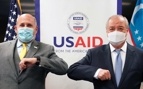 Two men in business suits are performing an elbow bump greeting in front of a USAID (United States Agency for International Development) banner. Both individuals are wearing face masks, consistent with COVID-19 era health protocols. The setting appears to be an official diplomatic or aid-related meeting, flanked by the American flag and what appears to be the flag of Uzbekistan.