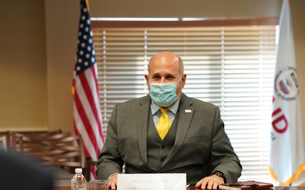 A man in a gray three-piece suit and yellow tie, wearing a light blue surgical face mask, is seated at a conference table in what appears to be an official government meeting room. Behind him are an American flag and a USAID organizational flag. A name placard on the table references 'USAID' and an official title, though the full text is partially obscured.
