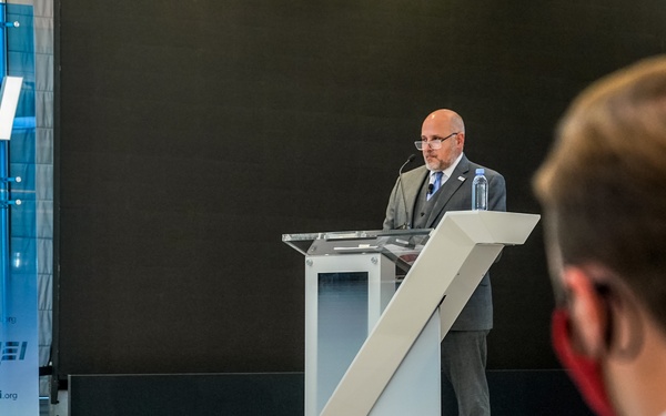 A bald man wearing glasses and a gray suit is speaking at a podium during a formal event hosted by AEI (American Enterprise Institute). The setting is a professional stage with branded signage displaying 'AEI' and the website 'aei.org'. An audience member in the foreground, wearing a face mask, is observing the presentation.