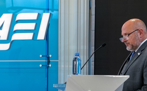 A bald man with a beard and glasses is standing at a white podium, appearing to read from notes or a prepared speech. The event is hosted by AEI (American Enterprise Institute), as indicated by the prominent blue branded backdrop behind him. The setting appears to be a formal conference or speaking event.