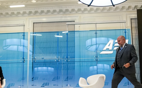 Two individuals are on a professional stage at an AEI (American Enterprise Institute) event. A woman is seated in a white modern chair on the left, while a man in a dark suit is walking toward the second empty white chair on the right. The stage features prominent AEI branding on backdrop panels and name placards are visible in front of the chairs.
