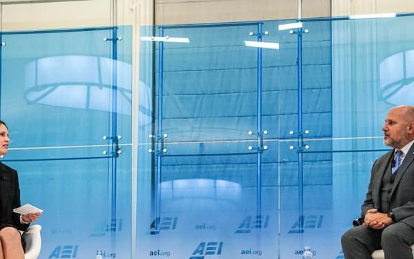 Two individuals are seated on a stage at an American Enterprise Institute (AEI) event, engaged in a formal discussion or interview. Name placards identify the speakers as Katherine Zimmerman (left) and John Barsa (right). The setting appears to be a professional policy forum with AEI branding prominently displayed throughout the backdrop.