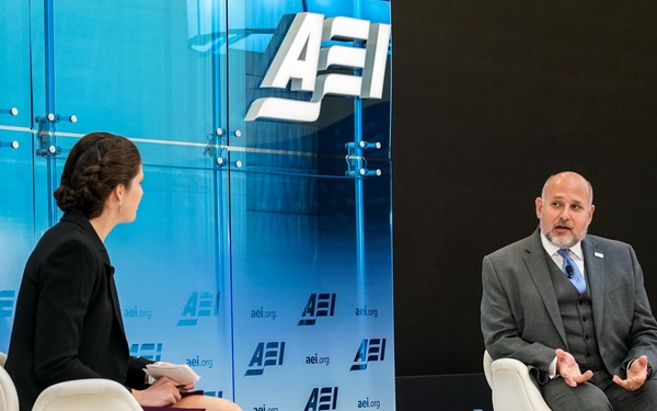 Two individuals are seated on a stage at an AEI (American Enterprise Institute) event, engaged in what appears to be a formal interview or panel discussion. A woman in a black blazer and purple skirt faces a man in a gray suit identified by a nameplate as 'John Barsa'. The setting features the AEI branding prominently displayed on a backdrop and glass wall.