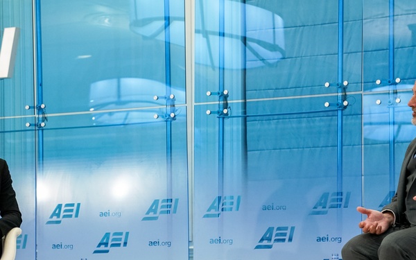 Two individuals are seated on a stage at an American Enterprise Institute (AEI) event, engaged in what appears to be a formal discussion or interview. Name placards identify the participants as Katherine Zimmerman (left) and John Barsa (right). The setting features a branded AEI backdrop with the organization's logo and website (aei.org) prominently displayed.