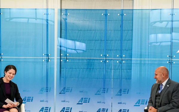 Two individuals are seated on a stage at an American Enterprise Institute (AEI) event, engaged in what appears to be a moderated discussion or interview. Name placards identify the participants as Katherine Zimmerman (left) and John Barsa (right). The setting features the AEI branding prominently displayed on backdrop panels and a repeating logo banner.