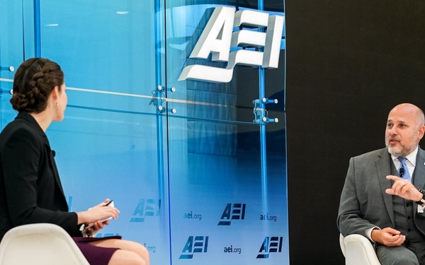Two individuals are seated on a stage at an AEI (American Enterprise Institute) event, engaged in what appears to be a formal interview or panel discussion. A woman in a black blazer and burgundy skirt faces a man identified by a nameplate as 'John Barsa', who is wearing a gray suit. The setting features prominent AEI branding on a glass backdrop and repeating logos on a banner.