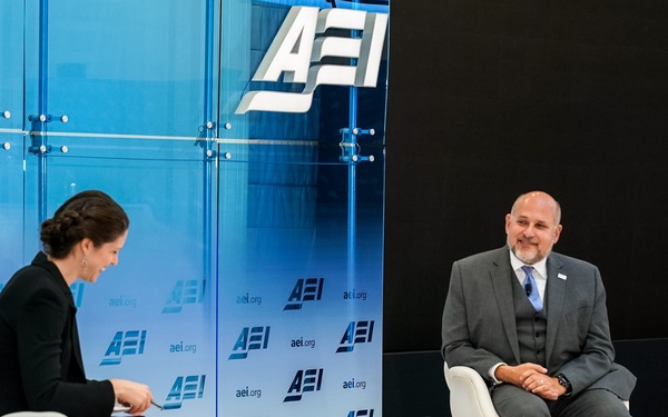 Two individuals are seated on a stage at an AEI (American Enterprise Institute) event, appearing to conduct an interview or panel discussion. A woman on the left holds papers or a notepad, while a man on the right, identified by a nameplate as 'John Barsa', sits in a relaxed posture. The setting features a professional backdrop with repeated AEI branding and aei.org logos.