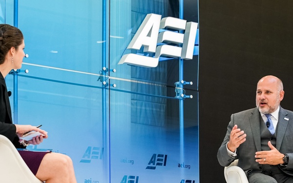 Two individuals are seated on a stage at an AEI (American Enterprise Institute) event, engaged in what appears to be a formal interview or panel discussion. A woman on the left holds a notepad and pen while facing a man identified by a nameplate as 'John Barsa', who is gesturing as he speaks. The backdrop prominently displays the AEI logo and website (aei.org) multiple times.