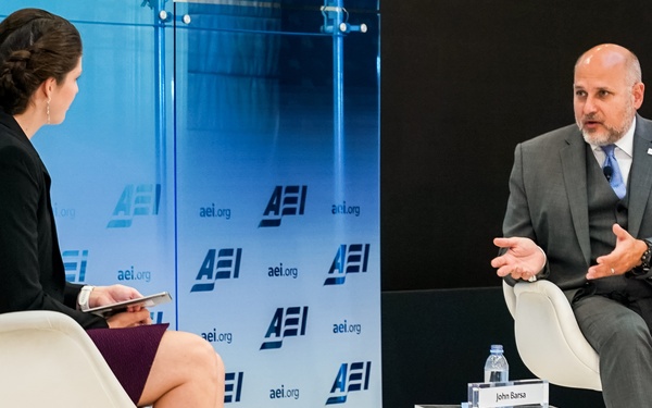 Two individuals are engaged in a formal interview or panel discussion at an AEI (American Enterprise Institute) event. A woman seated on the left holds a tablet and faces a man identified by a nameplate as 'John Barsa,' who is gesturing while speaking. The setting is a professional stage with a branded AEI backdrop displaying 'aei.org' repeatedly.