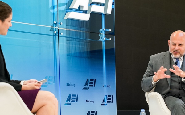 Two individuals are seated on a stage at an AEI (American Enterprise Institute) event, engaged in what appears to be a formal interview or panel discussion. A woman on the left holds a device and faces a man on the right, identified by a nameplate as John Barsa, who is speaking with animated hand gestures. The backdrop features repeated AEI branding and the website aei.org.