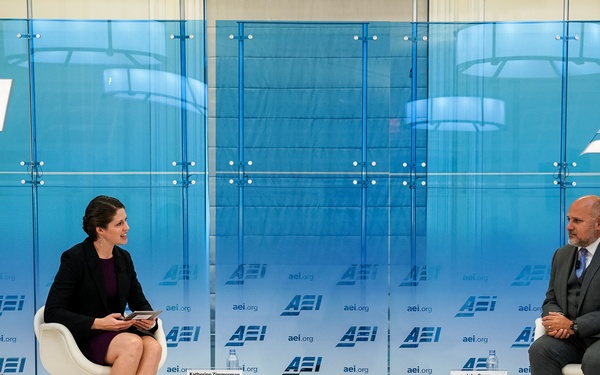 Two individuals are seated on a stage at an AEI (American Enterprise Institute) event, engaged in what appears to be a formal interview or panel discussion. Name placards identify the participants as Katherine Zimmerman (left) and John Barsa (right). The setting features a branded blue backdrop with repeated AEI logos and website URLs.