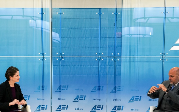 Two individuals are seated in white chairs on a stage at an AEI (American Enterprise Institute) event, engaged in a formal discussion or interview format. Name placards identify the participants as Katherine Zimmerman (left) and John Barsa (right). The setting features a branded blue backdrop with repeated AEI and aei.org logos.