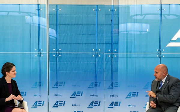 Two individuals are seated in white chairs on a stage at the American Enterprise Institute (AEI), engaged in a formal discussion or interview. Name placards identify the participants as Katherine Zimmerman (left) and John Barsa (right). The setting features AEI branding prominently displayed on blue glass panels and a step-and-repeat backdrop.