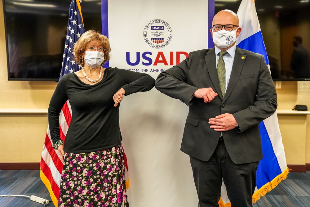 Two masked individuals are performing an elbow bump greeting in front of a USAID (United States Agency for International Development) banner. They are flanked by the American flag on the left and what appears to be the flag of El Salvador or a similar Central American nation on the right. The setting appears to be an official meeting or diplomatic event, likely during the COVID-19 pandemic period.