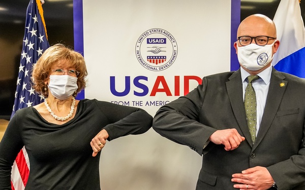 Two masked individuals are performing an elbow bump greeting in front of a USAID (United States Agency for International Development) banner. They are flanked by the American flag on the left and what appears to be the flag of El Salvador or a similar Central American nation on the right. The setting appears to be an official meeting or diplomatic event, likely during the COVID-19 pandemic period.