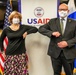Two masked individuals are performing an elbow bump greeting in front of a USAID (United States Agency for International Development) banner. They are flanked by the American flag on the left and what appears to be the flag of El Salvador or a similar Central American nation on the right. The setting appears to be an official meeting or diplomatic event, likely during the COVID-19 pandemic period.