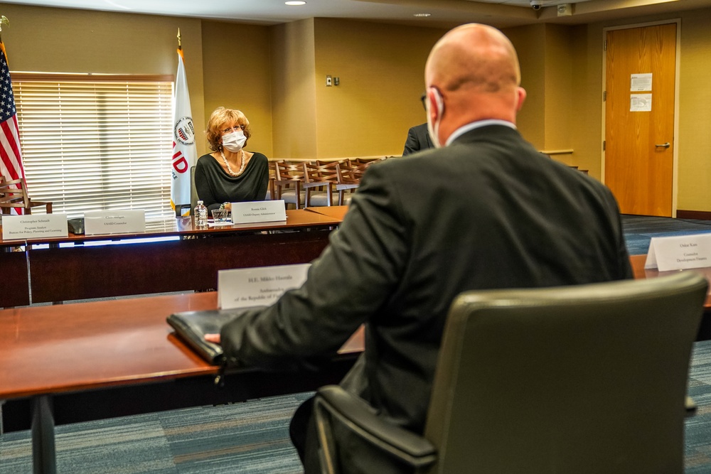 A formal meeting is taking place in a conference room, likely at a USAID facility, as indicated by the visible USAID flag and logo. A woman wearing a white face mask sits across the table from a bald man in a dark suit, whose back is to the camera. Name placards are visible on the table, suggesting an official diplomatic or governmental meeting.