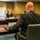 A formal meeting is taking place in a conference room, likely at a USAID facility, as indicated by the visible USAID flag and logo. A woman wearing a white face mask sits across the table from a bald man in a dark suit, whose back is to the camera. Name placards are visible on the table, suggesting an official diplomatic or governmental meeting.