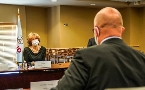 A formal meeting is taking place in a conference room, likely at a USAID facility, as indicated by the visible USAID flag and logo. A woman wearing a white face mask sits across the table from a bald man in a dark suit, whose back is to the camera. Name placards are visible on the table, suggesting an official diplomatic or governmental meeting.