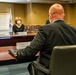 A formal meeting is taking place in a conference room, likely at a USAID facility, as indicated by the visible USAID flag and logo. A woman wearing a white face mask sits across the table from a bald man in a dark suit, whose back is to the camera. Name placards are visible on the table, suggesting an official diplomatic or governmental meeting.