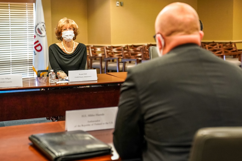 A formal meeting is taking place in a USAID conference room between officials seated at opposite sides of a table. The woman facing the camera is identified by her nameplate as Bonnie Glick, USAID Deputy Administrator, wearing a face mask. The man in the foreground, seen from behind, has a nameplate identifying him as H.E. Mikko Hautala, Ambassador of the Republic of Finland to the U.S.