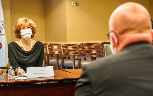 A formal meeting is taking place in a USAID conference room between officials seated at opposite sides of a table. The woman facing the camera is identified by her nameplate as Bonnie Glick, USAID Deputy Administrator, wearing a face mask. The man in the foreground, seen from behind, has a nameplate identifying him as H.E. Mikko Hautala, Ambassador of the Republic of Finland to the U.S.