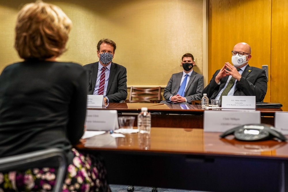 A formal meeting is taking place in a conference room with participants wearing face masks, suggesting a COVID-19 era setting. Three individuals sit on one side of a wooden conference table facing a fourth person whose back is to the camera. Name placards are visible on the table, with one legible card reading 'H.E. Mikko Hautala, Ambassador of the Republic of Finland to the U.S.'