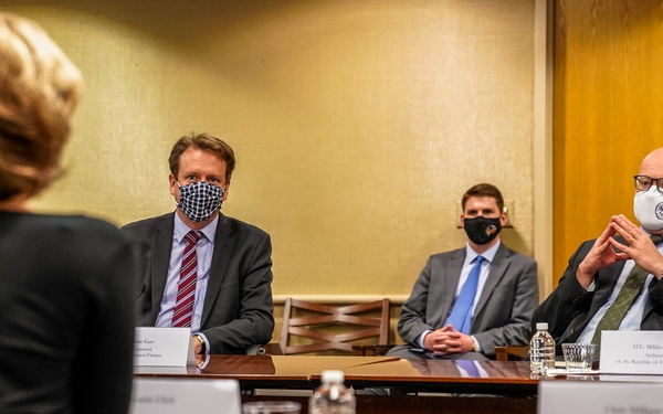 A formal meeting is taking place in a conference room with participants wearing face masks, suggesting a COVID-19 era setting. Three individuals sit on one side of a wooden conference table facing a fourth person whose back is to the camera. Name placards are visible on the table, with one legible card reading 'H.E. Mikko Hautala, Ambassador of the Republic of Finland to the U.S.'
