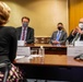 A formal meeting is taking place in a conference room with participants wearing face masks, suggesting a COVID-19 era setting. Three individuals sit on one side of a wooden conference table facing a fourth person whose back is to the camera. Name placards are visible on the table, with one legible card reading 'H.E. Mikko Hautala, Ambassador of the Republic of Finland to the U.S.'